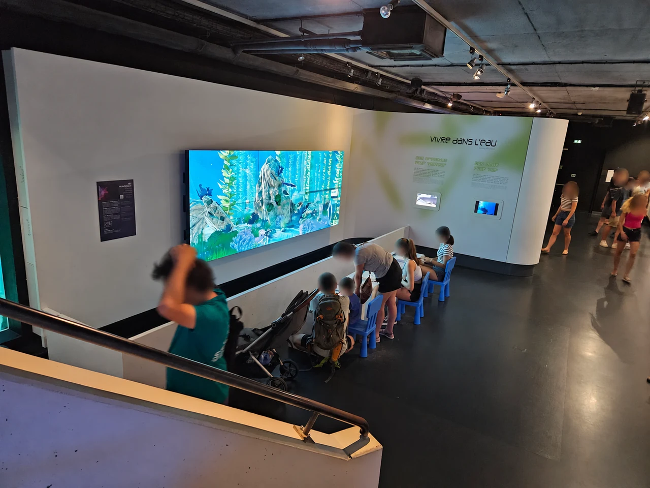 Visiteurs assis face au mur d’écrans de l’aquarium virtuel, vus depuis l’escalier