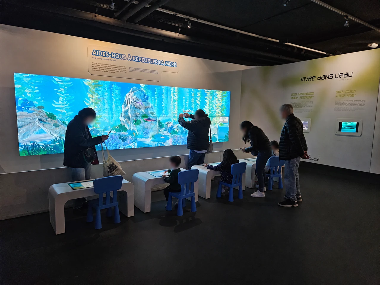Visiteurs, dont des enfants, devant le mur d’écrans de l’aquarium virtuel, avec les tables tactiles au premier plan