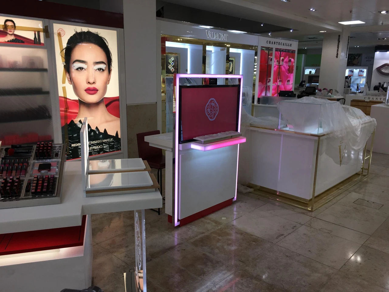 Espace de vente de produits de beauté avec une borne à grand écran rose encadrée de lumière, devant des affiches de maquillage