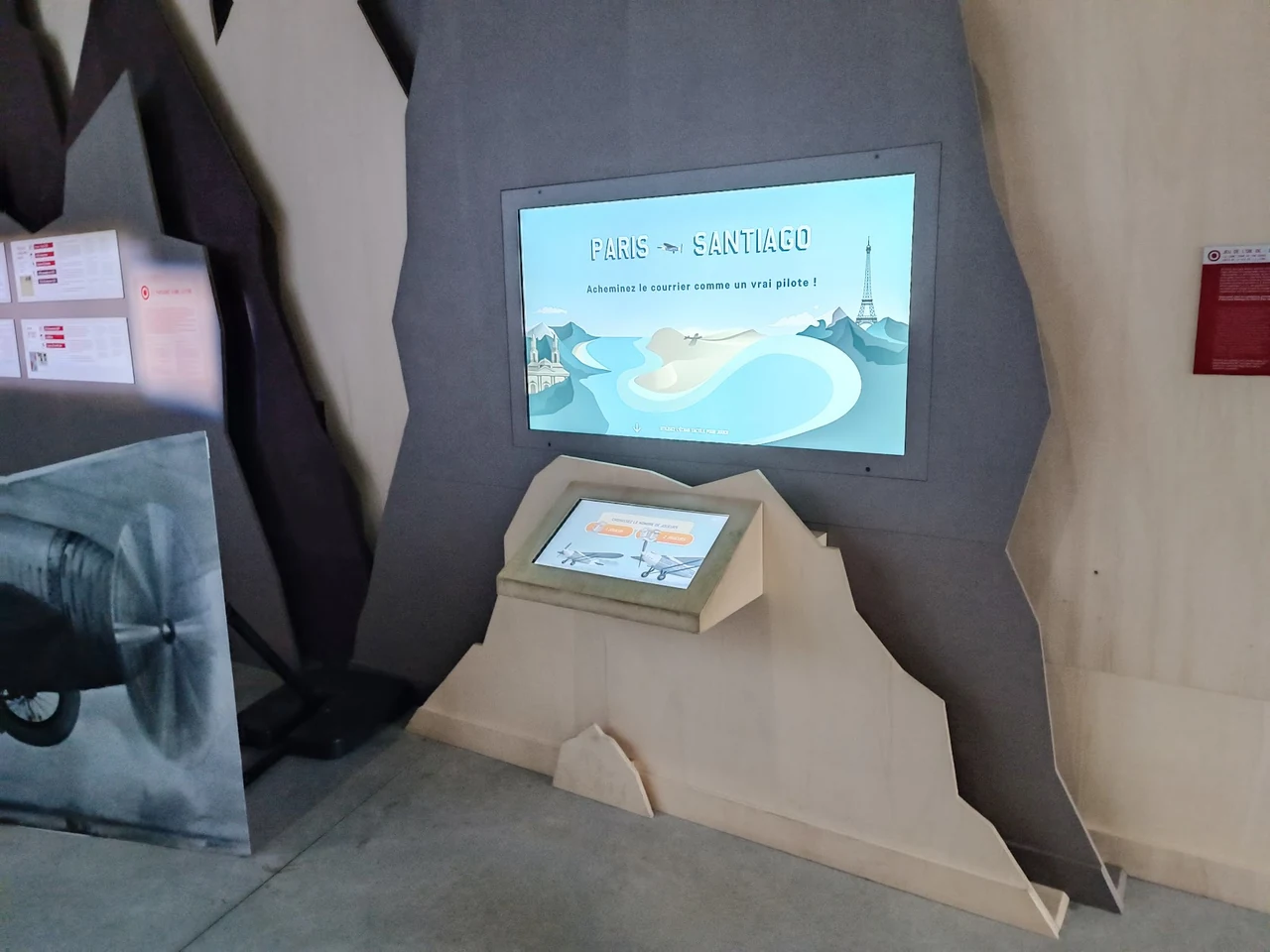 Borne tactile inclinée posée sur un décor en bois découpé, sous un écran mural
