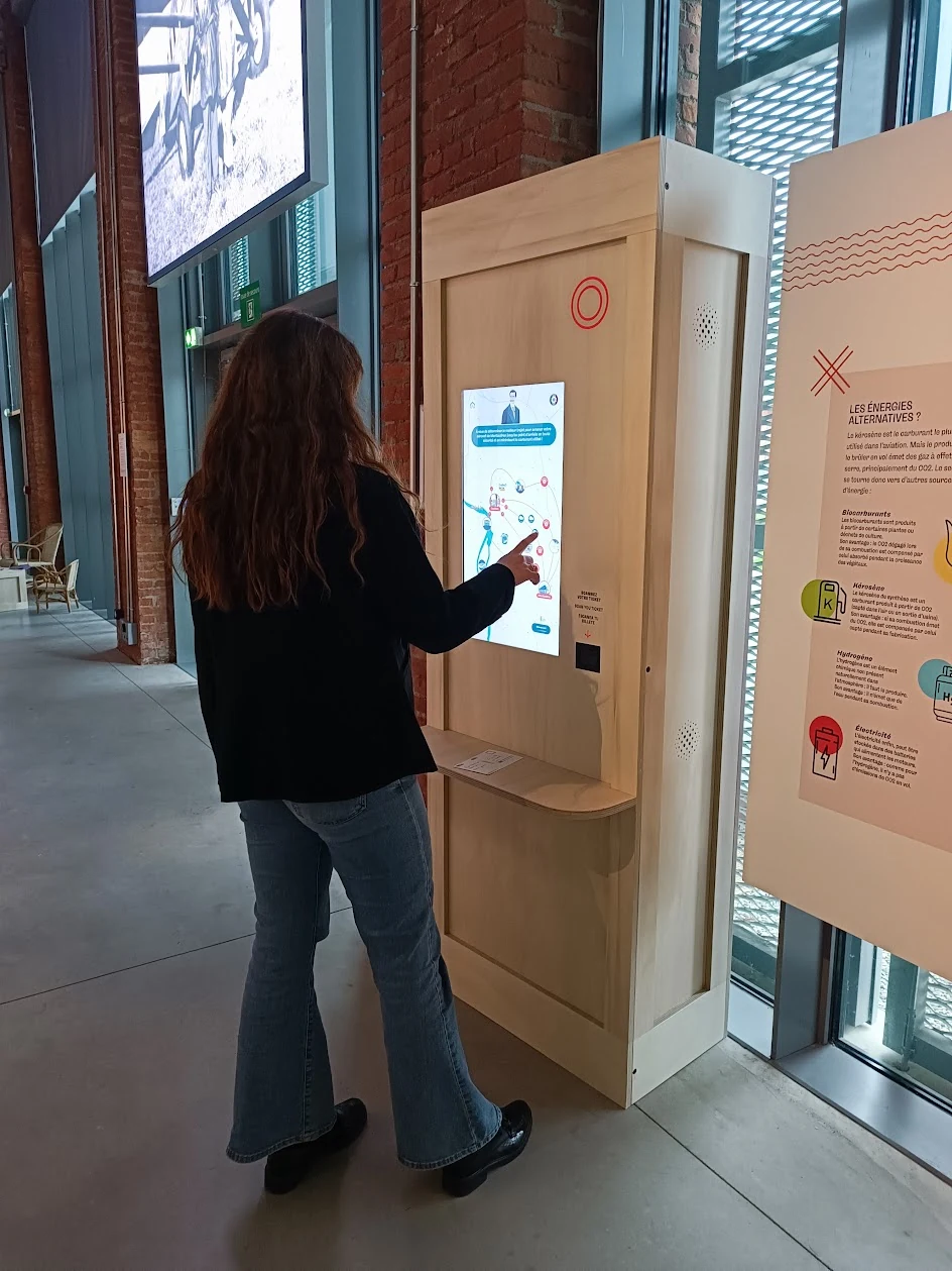Visiteuse touchant une borne tactile en bois dans un couloir vitré du musée