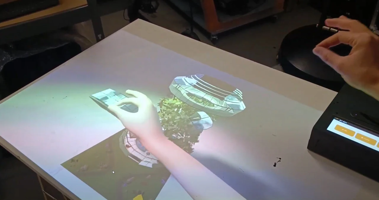 Main touchant une image projetée sur la table par la lampe interactive EY, à côté d’un petit boîtier à écran