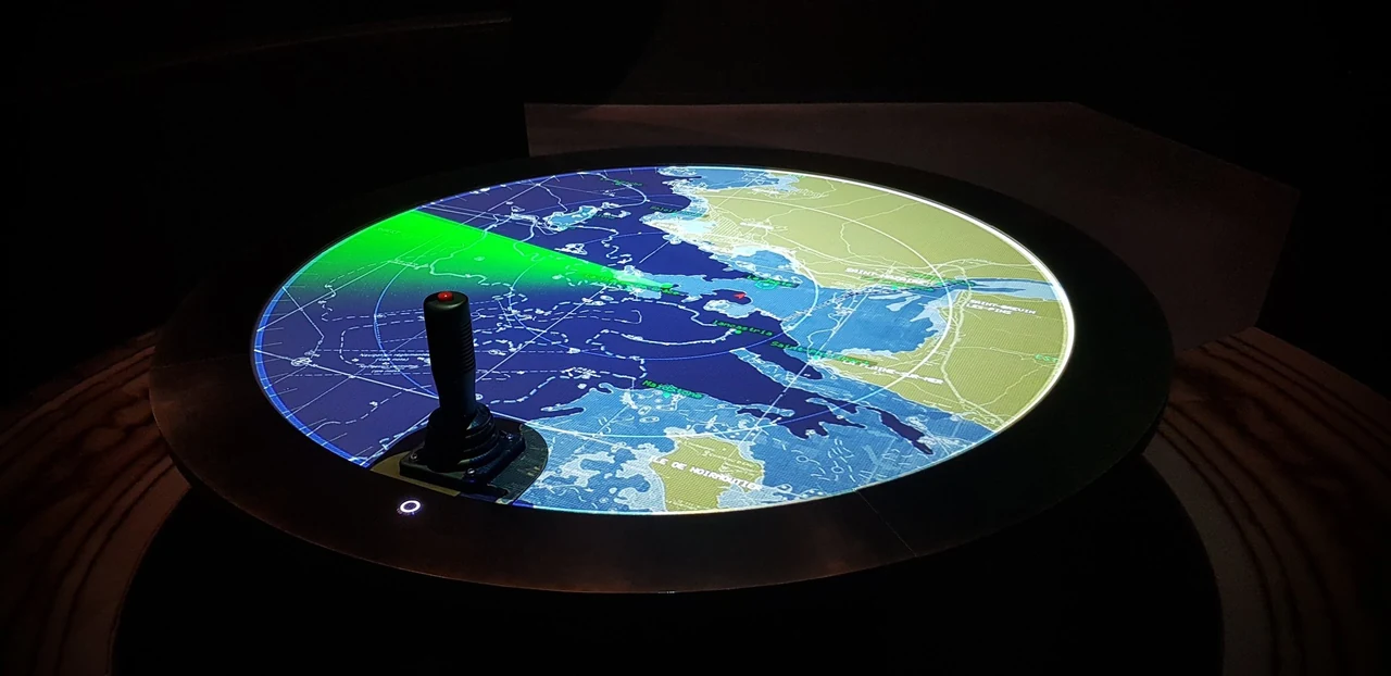 Table ronde projetant une carte marine avec un secteur radar vert, et son joystick, dans une salle sombre