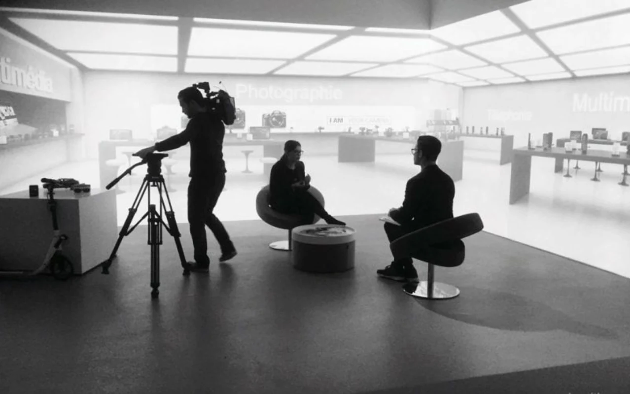 Photographie en noir et blanc : une personne près d’une caméra sur trépied et deux personnes assises devant un décor projeté