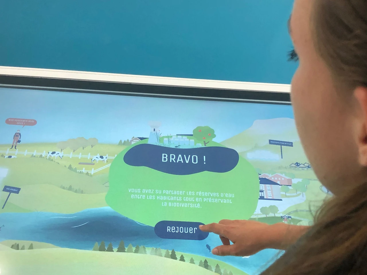 Personne de profil touchant le bouton « Rejouer » du message « Bravo ! » du jeu sur la gestion de l’eau, sur une grande table tactile