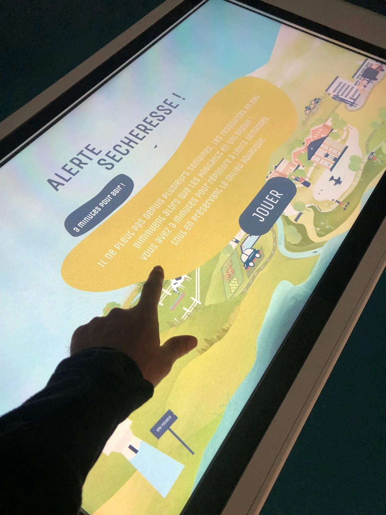 Main touchant le message « Alerte sécheresse ! » du jeu, sur la carte illustrée de la table tactile