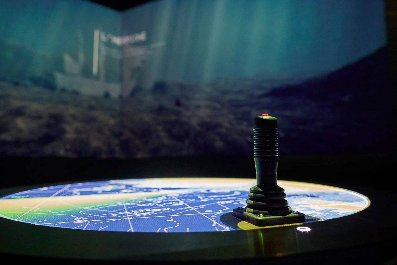 Joystick posé sur une table ronde projetant une carte marine, devant une vue sous-marine projetée sur l’écran courbe