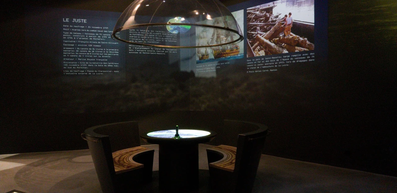 Salle sombre avec deux fauteuils autour de la table centrale lumineuse, face à un panneau d’exposition