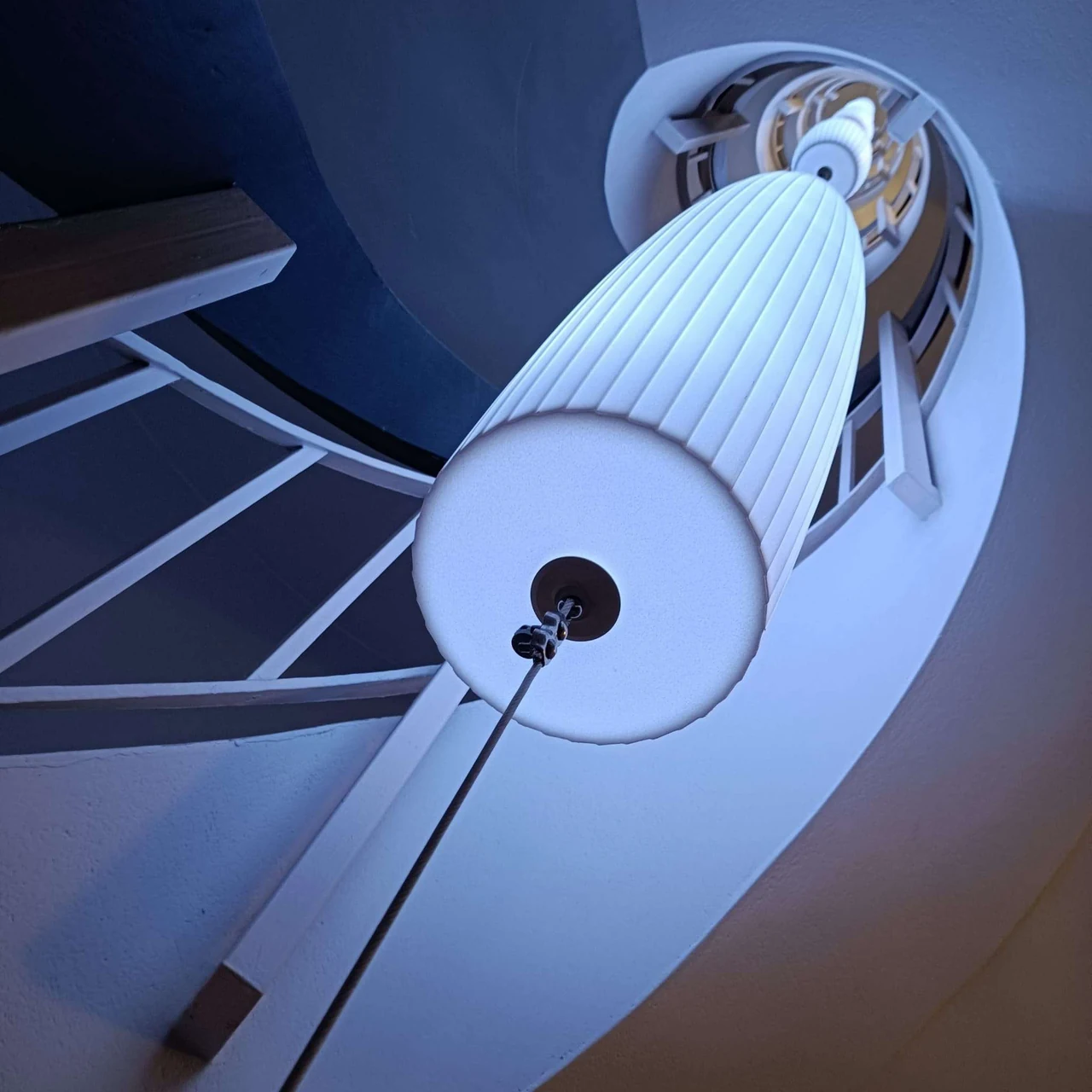 Lustre cylindrique blanc suspendu dans une cage d’escalier en spirale, vu d’en dessous, éclairé de bleu